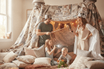 Familie im hellen Wohnzimmer baut eine magische Kissenburg aus Decken, Kissen und Lichterketten.